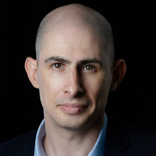Photo of Jason Steinhauer looking serious and staring at the camera. His face is half in shadow. He's wearing a blue shirt and his hair is shaved bald.